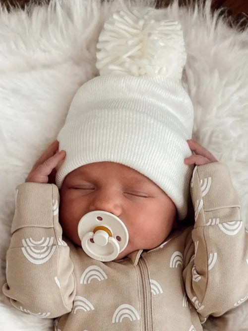Newborn baby sleeping with a white pom-pom hat and pacifier, wearing a tan onesie with rainbow designs.