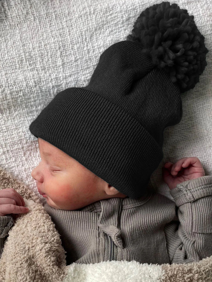 Newborn baby resting in a cozy blanket, wearing a black beanie with a pom-pom on top.