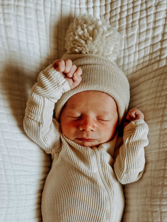 Newborn baby in cream outfit and pom-pom hat, peacefully sleeping on a textured white blanket.