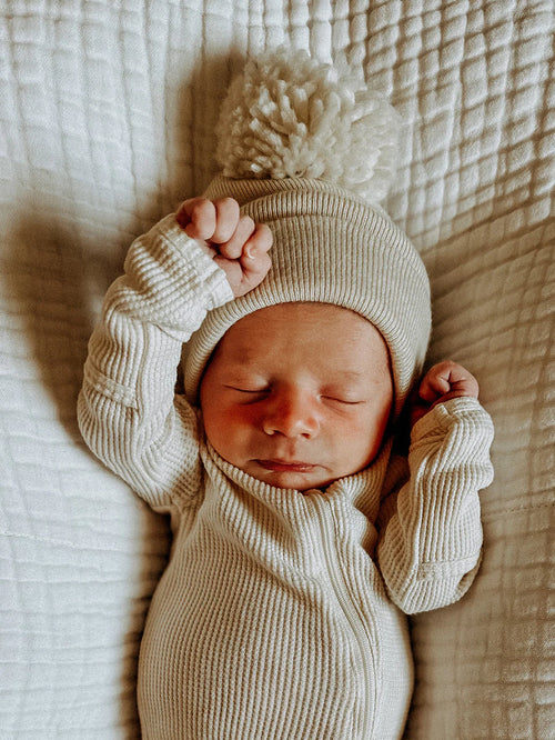 Newborn baby in cream outfit and pom-pom hat, peacefully sleeping on a textured white blanket.