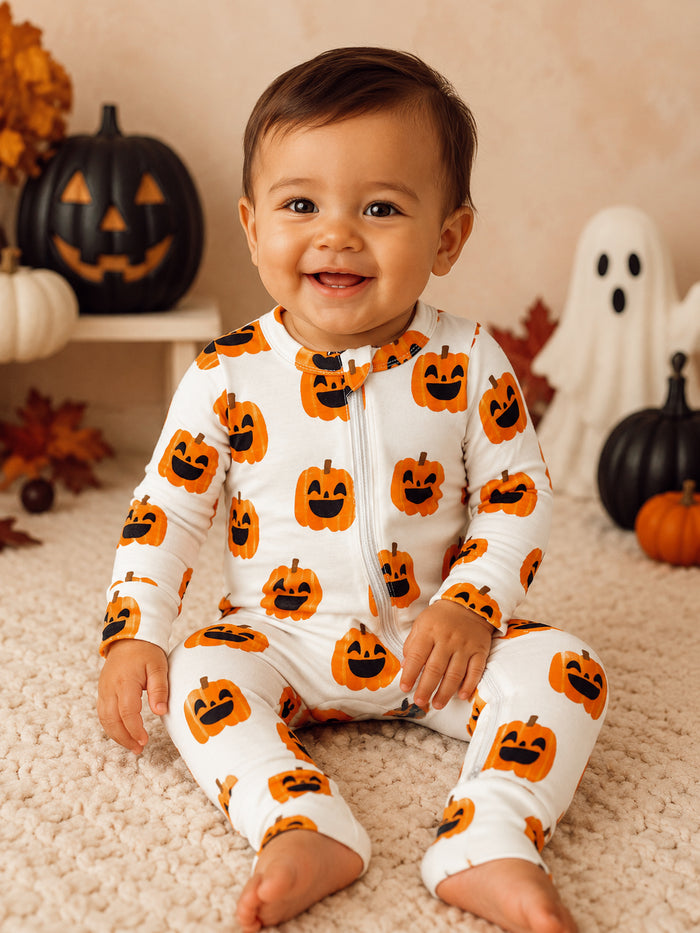 Smiling baby wearing pumpkin-print pajamas, surrounded by fall decorations and playful Halloween elements.