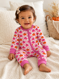 Smiling toddler in a pink pumpkin-patterned onesie sitting on a cozy blanket, surrounded by autumn decor.