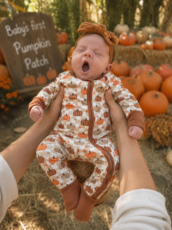 Baby in pumpkin-themed outfit yawning, held up in front of a "Baby's first Pumpkin Patch" sign and pumpkins.