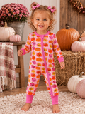 Smiling toddler in pumpkin-patterned pajamas stands on a cozy rug with pumpkins and flowers in the background.
