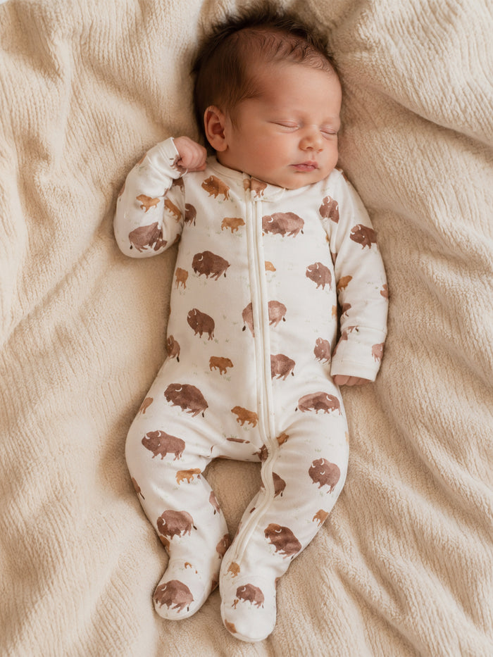 Infant asleep on a soft blanket, wearing a zip-up onesie with a bison pattern in earthy tones.