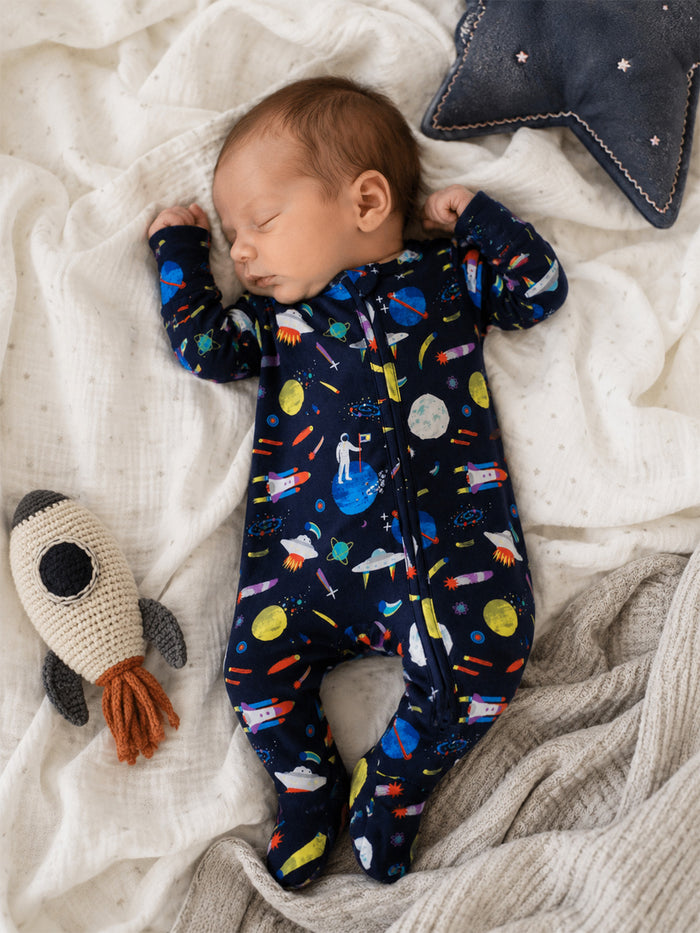 Sleeping baby in a space-themed onesie, surrounded by a blanket and plush rocket toy.