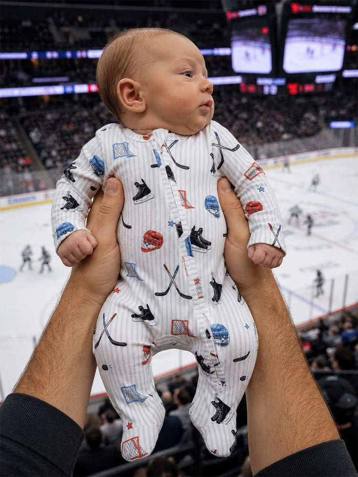 Baby in a sports-themed onesie held by an adult, with an ice hockey game in the background.