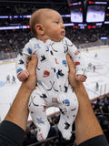 Baby in a sports-themed onesie held by an adult, with an ice hockey game in the background.