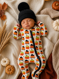 A sleeping baby in a pumpkin-themed onesie and black beanie, surrounded by autumn leaves and decorative pumpkins.