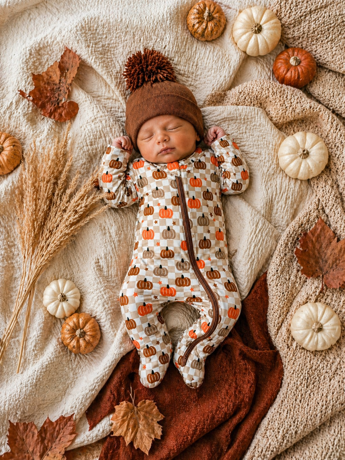 Sleeping baby dressed in a pumpkin-patterned onesie, surrounded by autumn leaves and pumpkins on a cozy blanket.