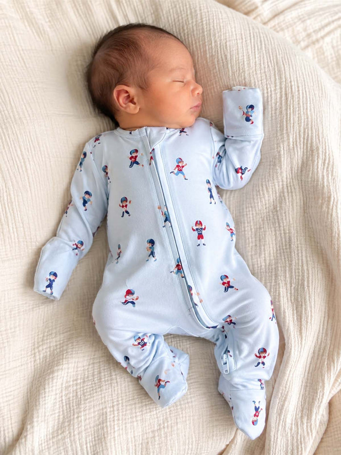 Sleeping baby in a blue onesie with sports-themed prints, lying on a textured light blanket.