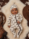 Smiling baby in horse-patterned outfit on soft fur with cowboy hat and boots nearby.