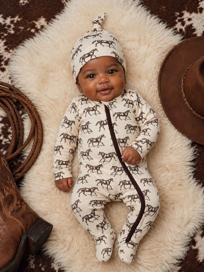 Smiling baby in horse-patterned outfit on soft fur with cowboy hat and boots nearby.