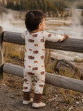Toddler in bison-printed pajamas, leaning on a wooden fence, gazing at a natural landscape.