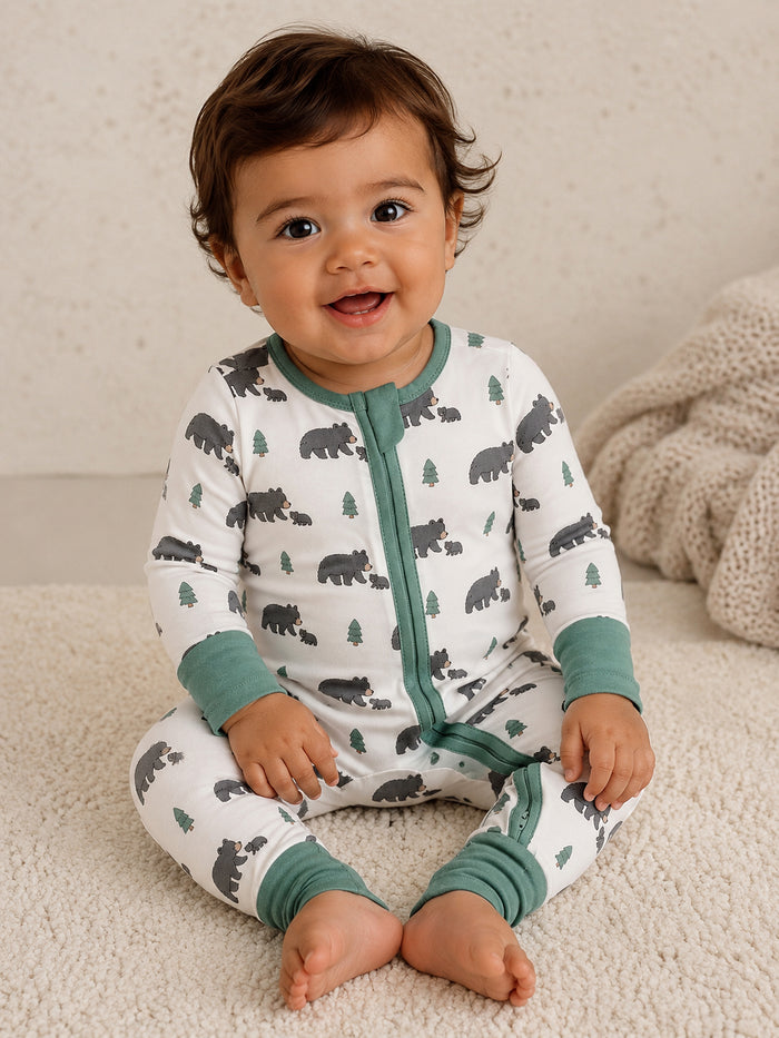 Smiling baby wearing bear-print pajamas, sitting on a soft cream rug with textured blankets in the background.