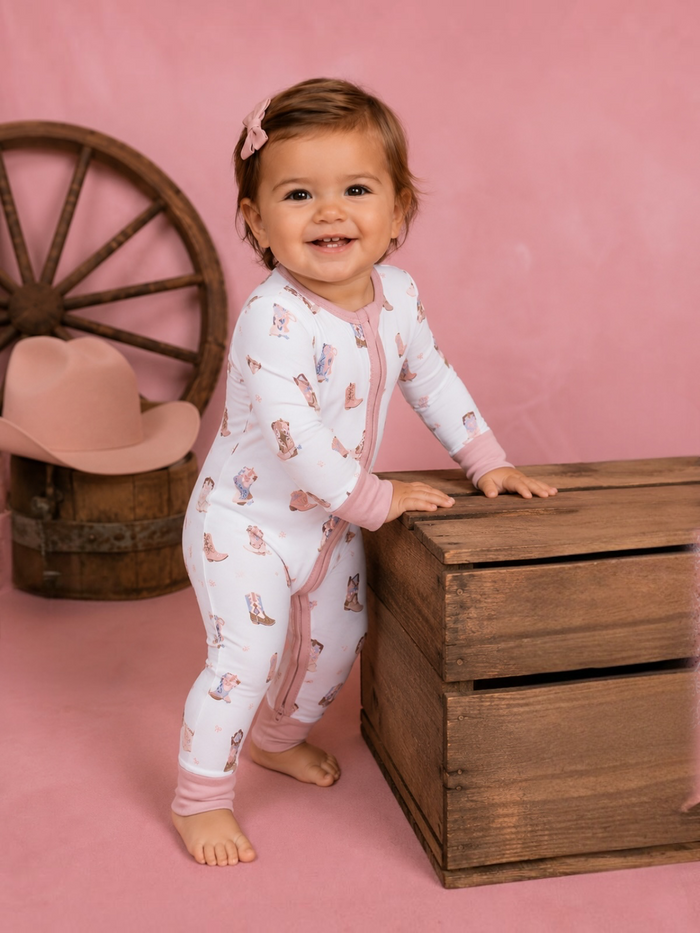 Smiling toddler in a patterned onesie stands by a wooden crate against a pink background.