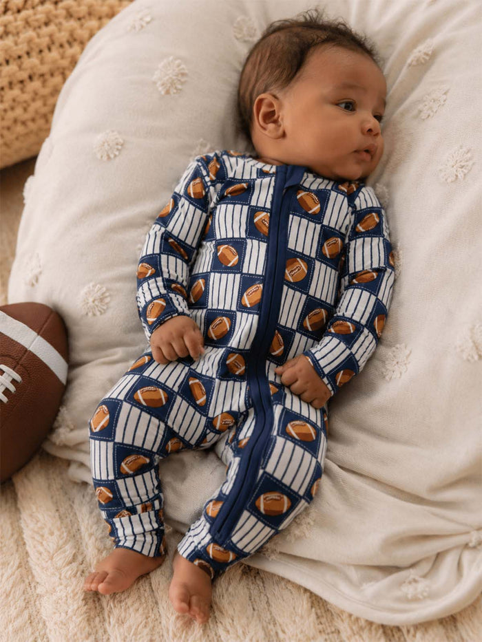 Baby in blue football-patterned onesie, lying on a soft blanket with a football nearby.
