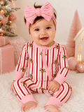 Smiling baby in festive striped pajamas with gingerbread design, pink headband, sitting on fluffy surface.