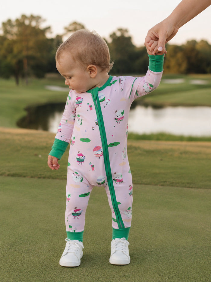 Toddler in colorful ice cream-themed onesie, holding an adult's hand on a green outdoor surface.