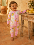 Smiling toddler in a lemon-patterned onesie stands by a table with lemons, against a rustic background.