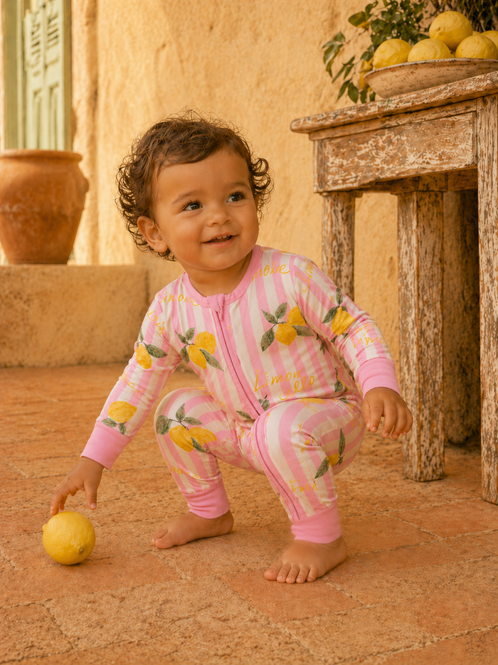 Smiling child in lemon-patterned pajamas squatting beside a lemon on a rustic outdoor surface.