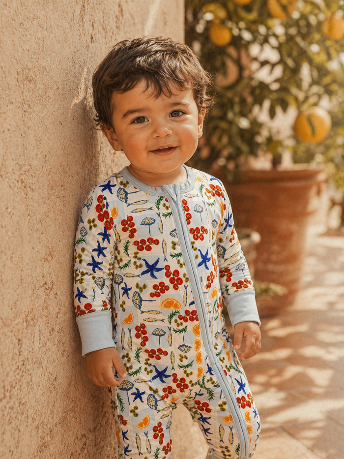 Smiling toddler in colorful patterned pajamas stands against a wall with greenery in the background.