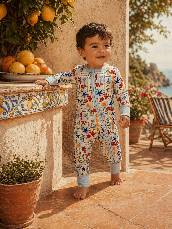 Smiling toddler in colorful onesie near fruit bowl, surrounded by plants and scenic outdoor setting.