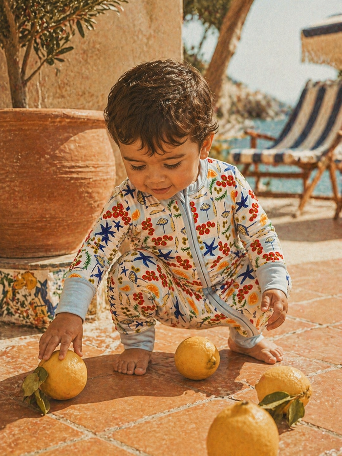 Child in colorful patterned outfit squatting, reaching for lemons on a sunny patio with a sea view.