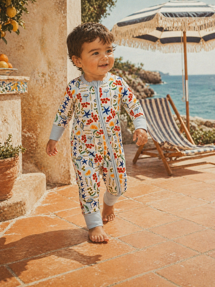 Smiling child in colorful pajamas walking outdoors by the sea, with a beach umbrella and chair in the background.