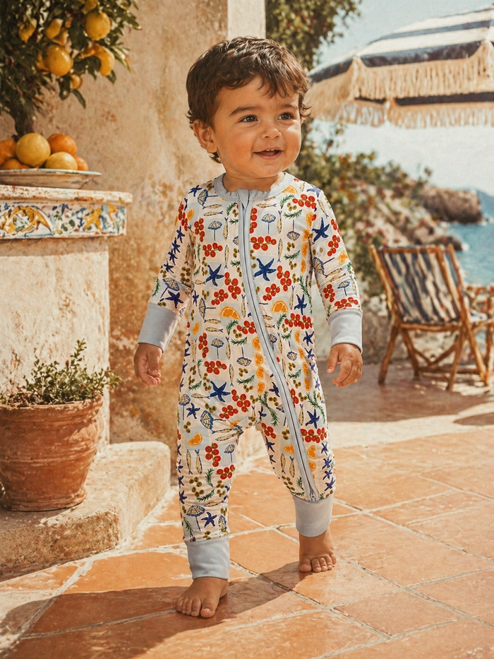 Toddler in a colorful, floral-patterned onesie smiles while walking on a sunny patio with a coastal view.