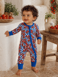Smiling toddler in colorful patterned onesie stands beside a table with tomatoes and herbs. Natural indoor setting.