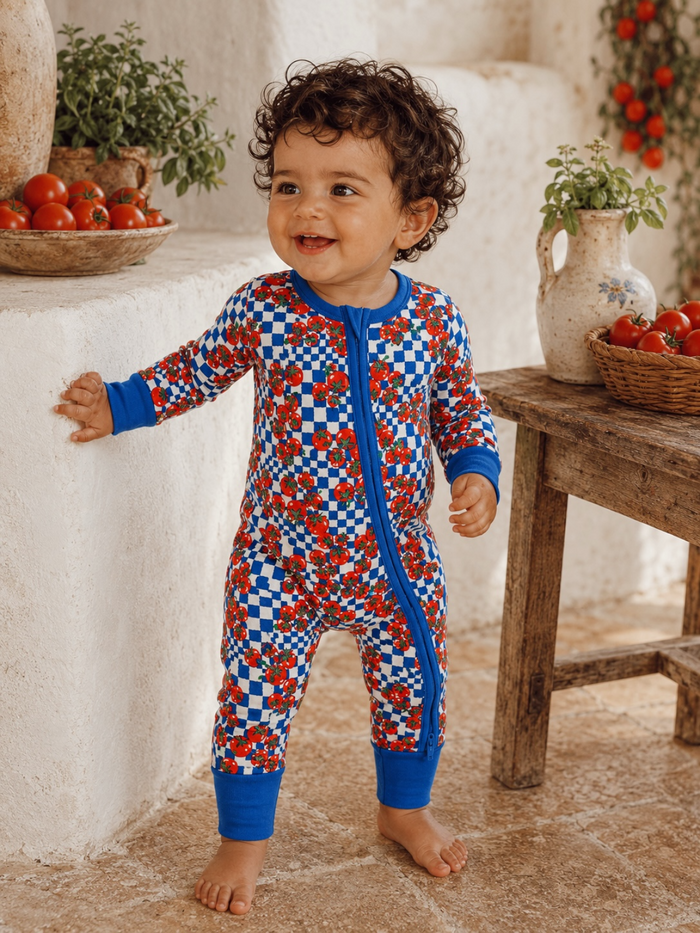 Smiling toddler in colorful patterned onesie stands beside a table with tomatoes and herbs. Natural indoor setting.