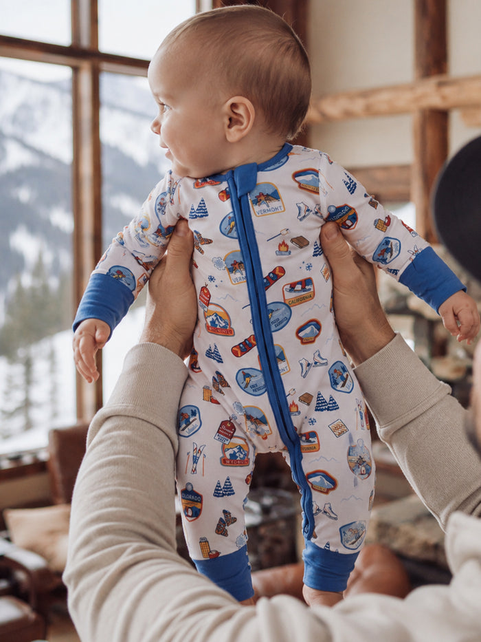 Baby in colorful adventure-themed onesie, held by an adult in a cozy cabin setting with mountain view.