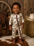 Smiling baby in horse-printed pajamas, standing on a cowhide rug with rustic decor in the background.