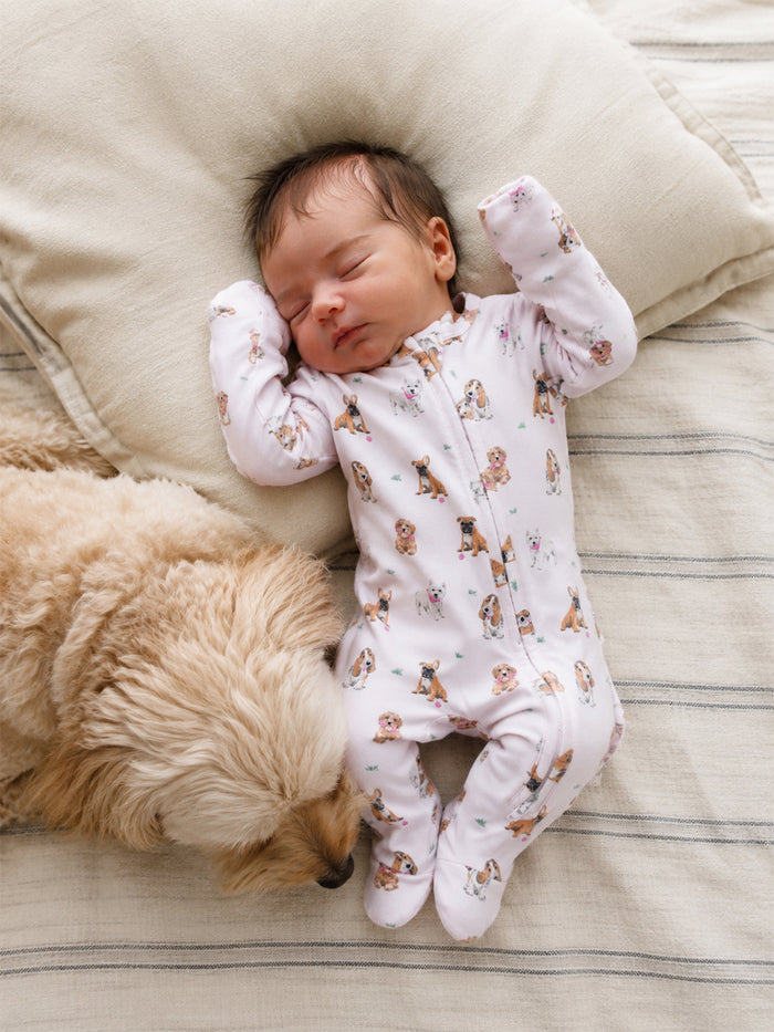 A sleeping baby in a dog-print onesie rests beside a fluffy dog on a cozy blanket.