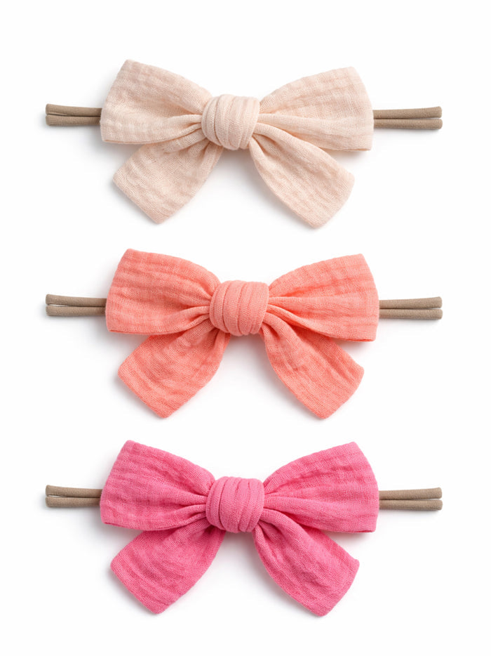 Three fabric hair bows in soft colors: cream, coral, and pink, attached to elastic bands.
