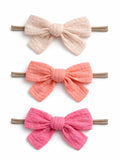 Three fabric hair bows in soft colors: cream, coral, and pink, attached to elastic bands.