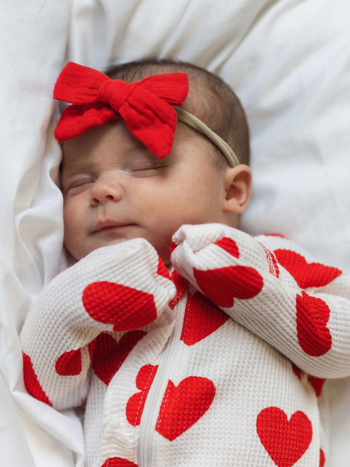 Newborn baby sleeping peacefully in a heart-patterned outfit with a red bow on a soft white blanket.
