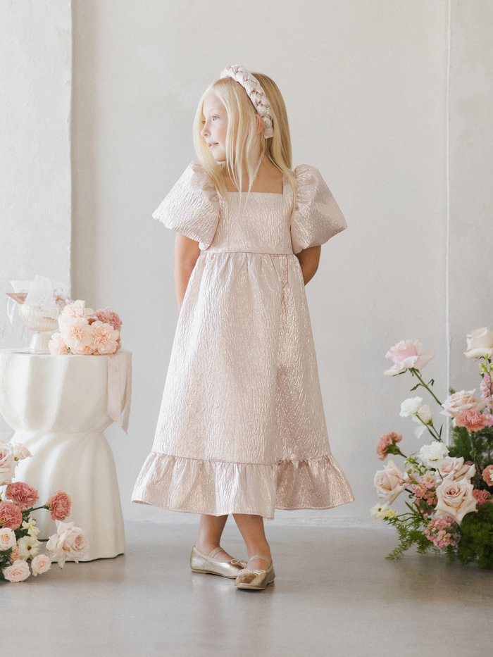 A girl in a pink dress with puffy sleeves stands against a light wall, surrounded by flowers.