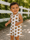 Smiling toddler in a bear-print outfit, playing outdoors near a white fence and gravel path, surrounded by greenery.