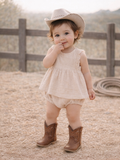 Toddler in a beige dress and cowboy hat, smiling while standing on straw-covered ground near a wooden fence.