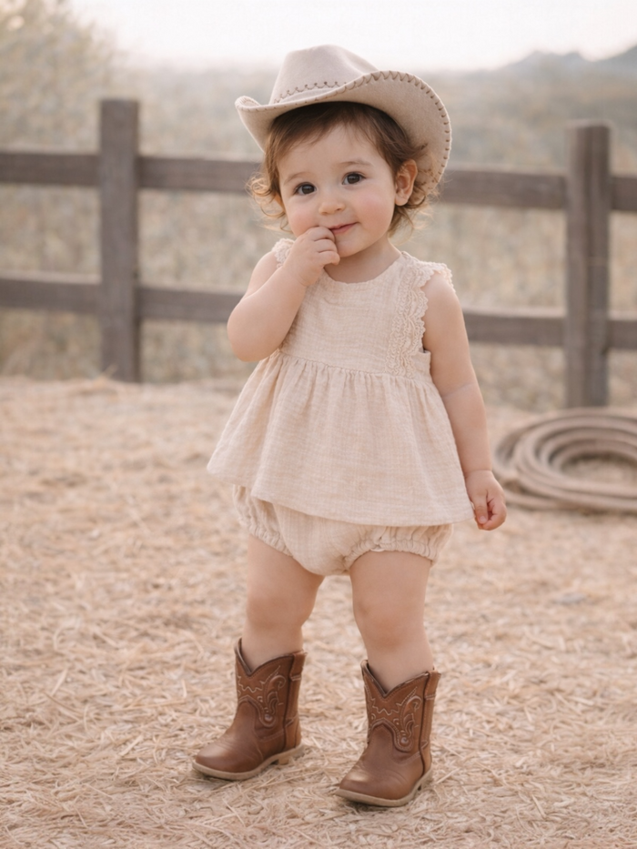 Toddler in a beige dress and cowboy hat, smiling while standing on straw-covered ground near a wooden fence.