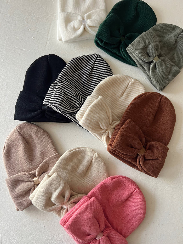 A variety of knitted beanies with bows in multiple colors arranged on a light background.