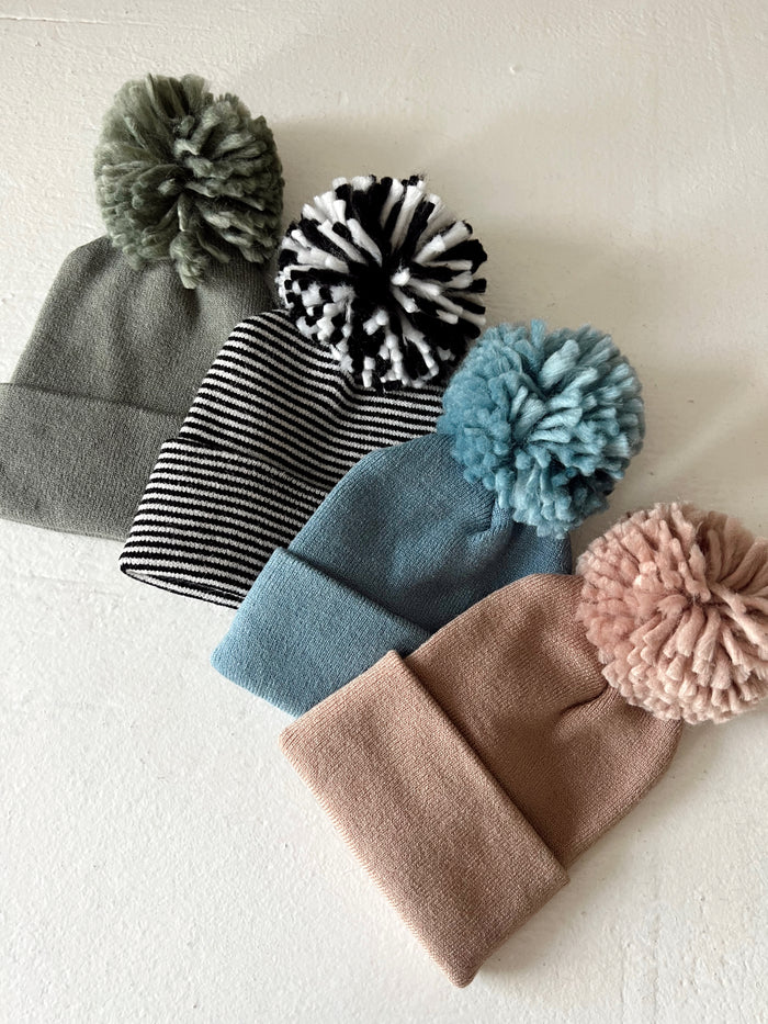 Four cozy beanies in various colors and patterns, featuring large pom-poms on top, displayed on a light background.