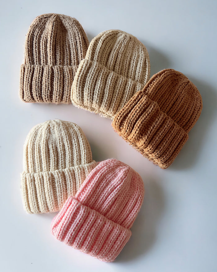 Five knitted beanies in cream, pink, light brown, and dark brown arranged on a white surface.