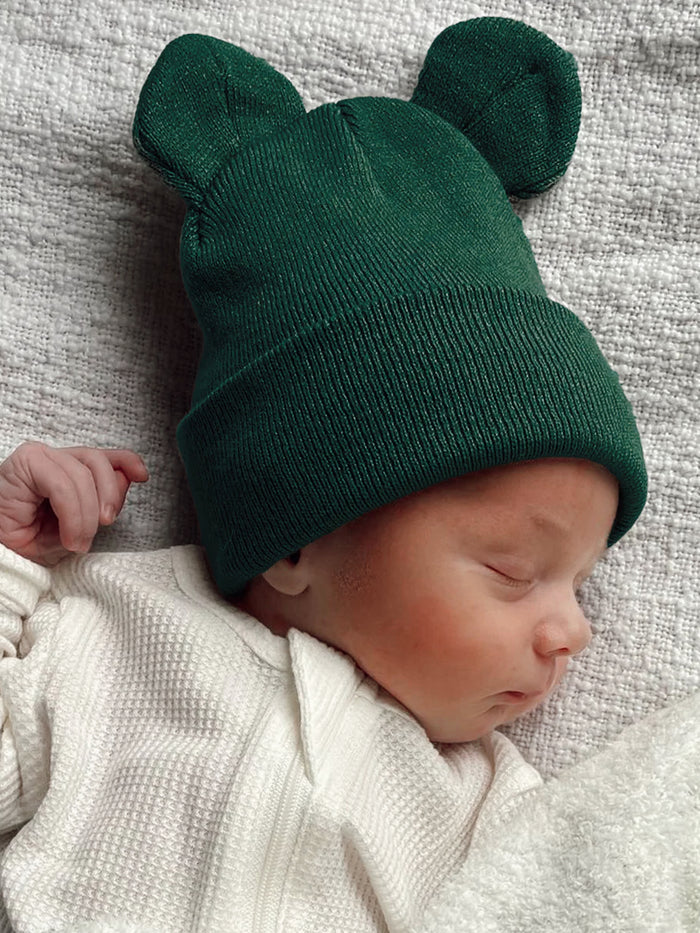 Baby sleeping in a white onesie, wearing a green knitted hat with ears, on a textured gray blanket.