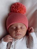 Baby sleeping peacefully in a pink pompom hat, resting on a soft blanket.