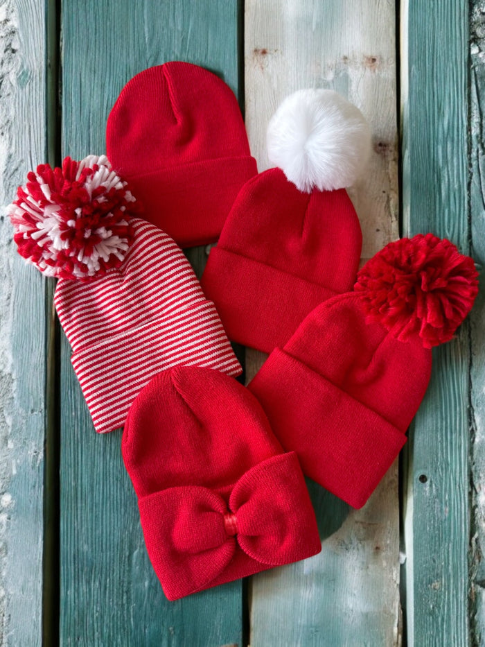 A collection of six red winter hats, including styles with pompoms and a bow, laid on a wooden surface.