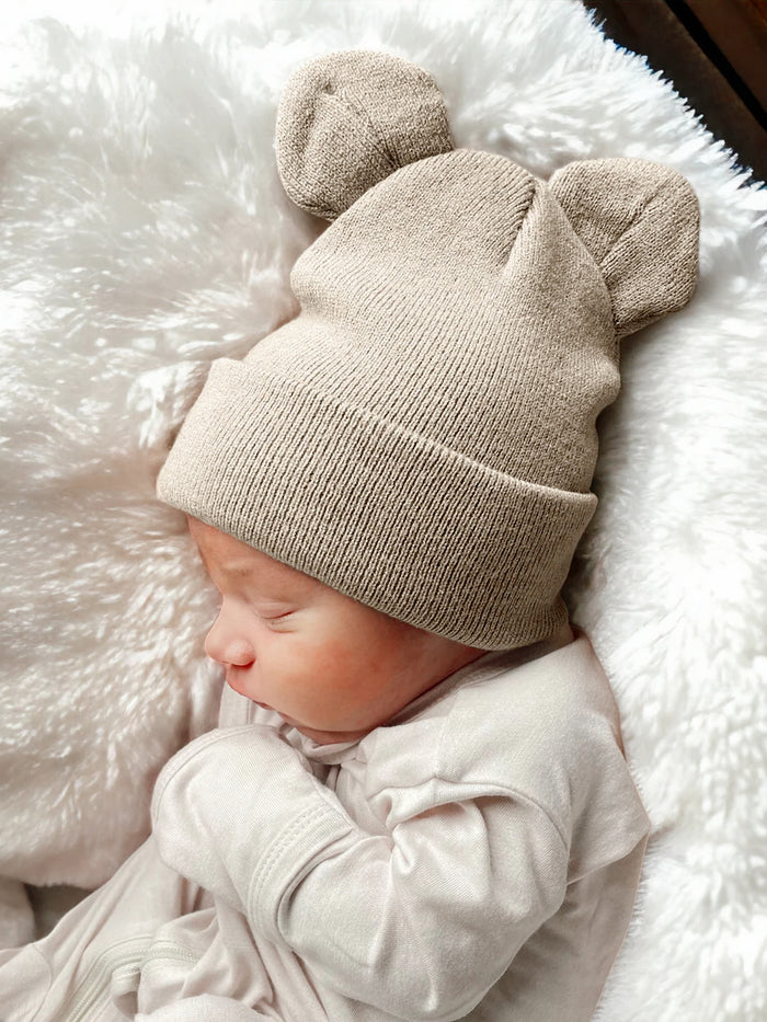 Newborn baby sleeping on a soft blanket, wearing a beige bear-ear knit hat.