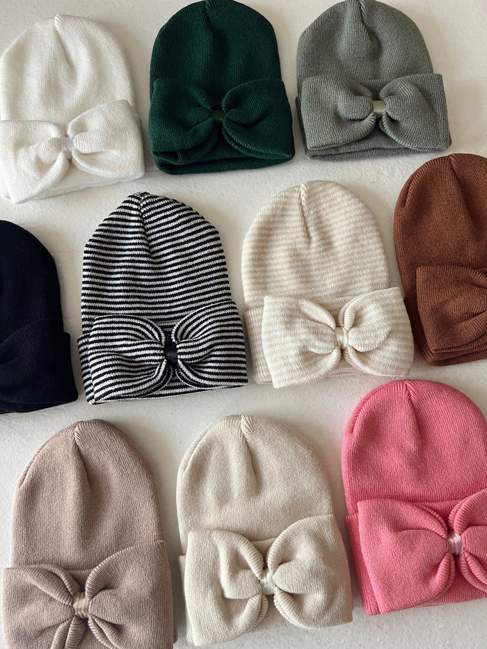 Colorful collection of beanies with bows, displayed in various styles and patterns on a white background.